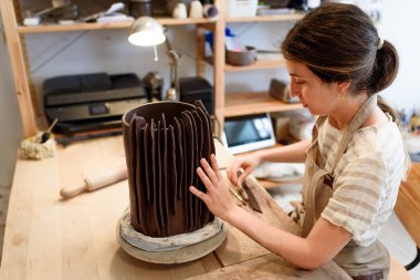 Çömlek atölyesinde çiğ seramik vazo ve kilden esrar yapan bir kadın. Doğal ışık kazıma, düzleştirme, şekillendirme ve yontma ile çömlek atölyesi. Kil tenceresi yapmak
