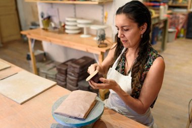 Geleceğin seramik ürünleri için çiğ seramik ve kil üreten genç bir kadın. Doğal ışık kazıma, düzleştirme, şekillendirme ve yontma ile çömlek atölyesi. Kil tenceresi yapmak