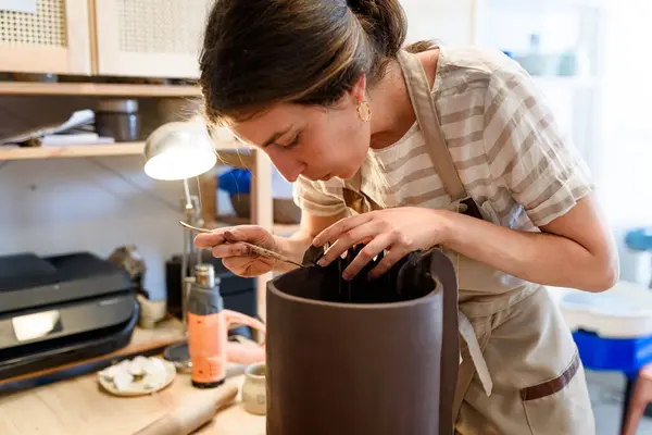 stock image woman in pottery workshop working raw ceramic vase and clay to make pot. Pottery workshop with natural light scraping, smoothing, shaping and sculpting. making clay pot