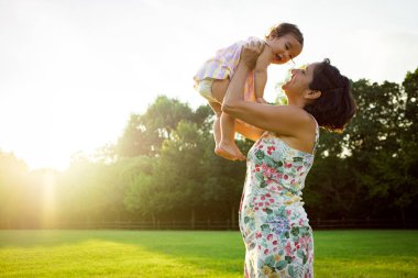 Anne, gün batımında mutlu bebek kızını doğada parkta havada tutuyor. Annelik ve annelikte uyum kavramı, birlikte mutlu anları paylaşıyor..