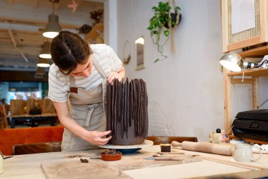 Çömlek atölyesinde çiğ seramik vazo ve kilden esrar yapan bir kadın. Doğal ışık kazıma, düzleştirme, şekillendirme ve yontma ile çömlek atölyesi. Kil tenceresi yapmak