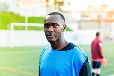 Genç siyahi Afrikalı Amerikalı adamın bir futbol sahasında eğitildiği, kameraya bakan portresi. Futbol sahasında spor ve egzersiz yapan genç bir sporcu