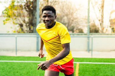 Futbol sahasında koşarken kamerada mutlu görünen gülümseyen siyah Afrikalı bir adamın portresi. Futbol sahasında spor ve egzersiz yapan genç bir sporcu