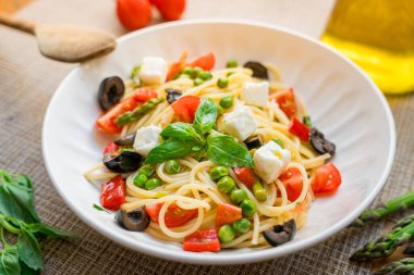 Makarna primavera spagettisi, vişneli domatesler, siyah zeytinler, feta peyniri ve fesleğen, ahşap masa üzerinde servis edilir, doğal ışık, italyan yemeği ve ev yapımı yemek.
