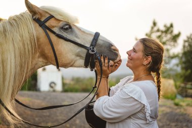 40 'larında bir kadın ahırda atına bakıyor, gün batımında gülümsüyor ve kaynaşıyor.