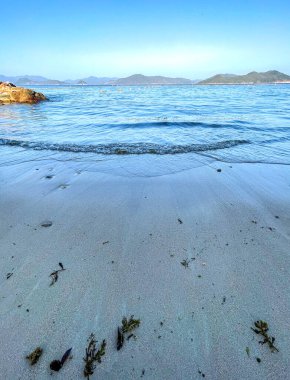 Açık deniz, su dalgaları, doğal taşlar, mavi gökyüzü ve dağlar.