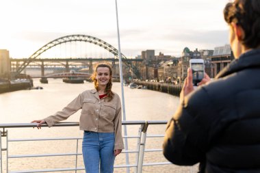 Genç bir çift günü Tyne 'da Newcastle' da birlikte geçiriyorlar. Kadın, erkek arkadaşının cep telefonuyla çektiği bir fotoğraf için poz veriyor. O sırada arkasında Tyne nehri olan bir köprüde duruyor..