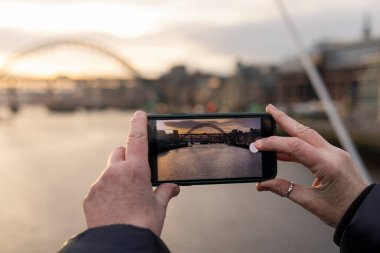 Tanımlanamayan bir kadın, Tyne nehrinin batımında cep telefonuyla fotoğraf çekiyor. Newcastle Upon Tyne 'da. Köprüler fotoğrafta görülebilir..