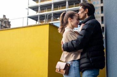 Duvarın karşısında yüz yüze duran genç bir çiftin yan görüntüsü, kucaklaşıyorlar ve öpüşüyorlar, birbirlerine aşıklar..