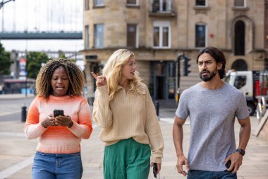 İngiltere, Newcastle 'da bir şehir sokağında duran üç yetişkin arkadaşın geniş açılı görüntüsü. Akıllı telefon kullanırken yol tarifi arıyorlar. Günlük giysiler giyiyorlar ve ne tarafa gideceklerini tartışıyorlar..