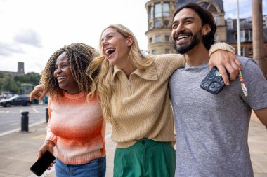 İngiltere, Newcastle 'da bir şehir sokağında duran üç yetişkin arkadaşın geniş açılı görüntüsü. Akıllı telefon kullanırken yol tarifi arıyorlar. Sıradan kıyafetler giyiyorlar ve kollarını birbirlerine dolayarak yürüyorlar..