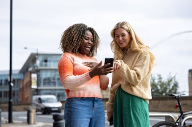 İngiltere 'nin Newcastle kentindeki bir caddede duran iki yetişkin kadının ön görüntüsü. Akıllı telefon kullanırken yol tarifi arıyorlar. Günlük giysiler giyiyorlar ve ne tarafa gideceklerini tartışıyorlar..