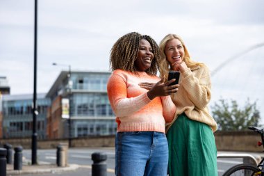 İngiltere 'nin Newcastle kentindeki bir caddede duran iki yetişkin kadının ön görüntüsü. Akıllı telefon kullanırken yol tarifi arıyorlar. Günlük giysiler giyiyorlar ve ne tarafa gideceklerini tartışıyorlar..