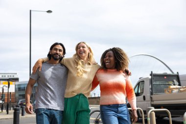 İngiltere, Newcastle 'da bir şehir caddesinde yürüyen üç yetişkin arkadaşın geniş açılı görüntüsü. Sıradan kıyafetler giyiyorlar ve kollarını birbirlerine dolayarak yürüyorlar..