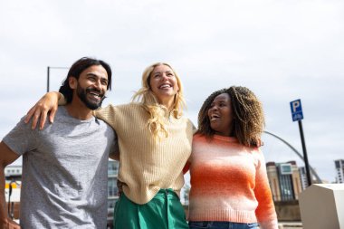 İngiltere, Newcastle 'da bir şehir caddesinde yürüyen üç yetişkin arkadaşın geniş açılı görüntüsü. Sıradan kıyafetler giyiyorlar ve kollarını birbirlerine dolayarak yürüyorlar..