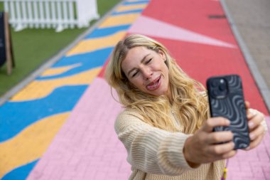 İngiltere, Newcastle 'da şehir merkezindeki bir caddede duran orta yaşlı bir kadının selfie çekimi. Günlük kıyafetler giyiyor, ayakta duruyor, gülümsüyor ve akıllı telefondan selfie çekerken kameraya bakıyor, dilini dışarı çıkarıyor..