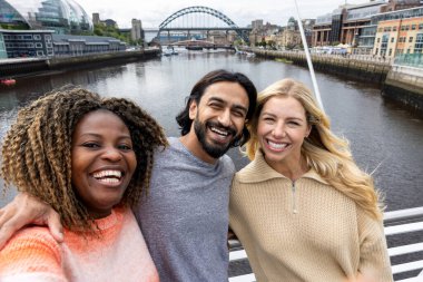 İngiltere, Newcastle 'da bir köprüden geçerken şehir merkezinde duran üç orta yaşlı arkadaşın selfie çekimi. Günlük giysiler giyiyorlar ve yan yana duruyorlar, gülümsüyorlar ve grup fotoğrafı çekerken kameraya bakıyorlar..