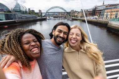 İngiltere, Newcastle 'da bir köprüden geçerken şehir merkezinde duran üç orta yaşlı arkadaşın selfie çekimi. Günlük giysiler giyiyorlar ve yan yana duruyorlar, gülümsüyorlar ve aptal bir grup selfie 'si çekerken kameraya bakıyorlar..