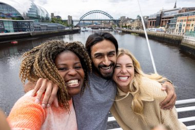 İngiltere, Newcastle 'da bir köprüden geçerken şehir merkezinde duran üç orta yaşlı arkadaşın selfie çekimi. Günlük giysiler giyiyorlar ve yan yana duruyorlar, gülümsüyorlar ve grup fotoğrafı çekerken kameraya bakıyorlar..