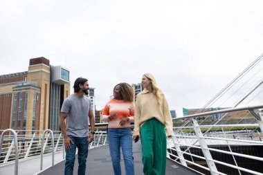 Üç yetişkin arkadaşın Newcastle, İngiltere 'de bir köprüden geçerken bir şehir caddesinde yürürken ön görüntüsü. Günlük giysiler giyiyorlar ve yan yana yürüyorlar, gülümsüyorlar ve sohbet ediyorlar..