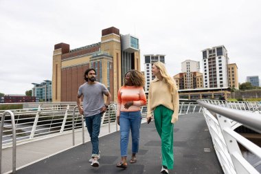 Üç yetişkin arkadaşın Newcastle, İngiltere 'de bir köprüden geçerken bir şehir caddesinde yürürken ön görüntüsü. Günlük giysiler giyiyorlar ve yan yana yürüyorlar, gülümsüyorlar ve sohbet ediyorlar..