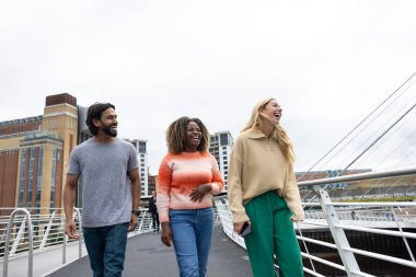 Üç yetişkin arkadaşın Newcastle, İngiltere 'de bir köprüden geçerken bir şehir caddesinde yürürken ön görüntüsü. Günlük giysiler giyiyorlar ve yan yana yürüyorlar, gülümsüyorlar ve sohbet ediyorlar..