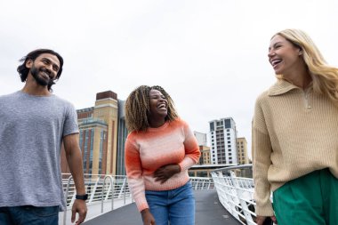 Üç yetişkin arkadaşın Newcastle, İngiltere 'de bir köprüden geçerken bir şehir caddesinde yürürken ön görüntüsü. Günlük giysiler giyiyorlar ve yan yana yürüyorlar, gülümsüyorlar ve sohbet ediyorlar..