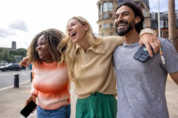 İngiltere, Newcastle 'da bir şehir sokağında duran üç yetişkin arkadaşın geniş açılı görüntüsü. Akıllı telefon kullanırken yol tarifi arıyorlar. Sıradan kıyafetler giyiyorlar ve kollarını birbirlerine dolayarak yürüyorlar..