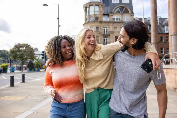 İngiltere, Newcastle 'da bir şehir sokağında duran üç yetişkin arkadaşın geniş açılı görüntüsü. Akıllı telefon kullanırken yol tarifi arıyorlar. Sıradan kıyafetler giyiyorlar ve kollarını birbirlerine dolayarak yürüyorlar..