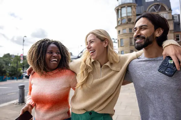 İngiltere, Newcastle 'da bir şehir sokağında duran üç yetişkin arkadaşın geniş açılı görüntüsü. Akıllı telefon kullanırken yol tarifi arıyorlar. Sıradan kıyafetler giyiyorlar ve kollarını birbirlerine dolayarak yürüyorlar..