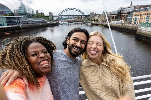 İngiltere, Newcastle 'da bir köprüden geçerken şehir merkezinde duran üç orta yaşlı arkadaşın selfie çekimi. Günlük giysiler giyiyorlar ve yan yana duruyorlar, gülümsüyorlar ve aptal bir grup selfie 'si çekerken kameraya bakıyorlar..