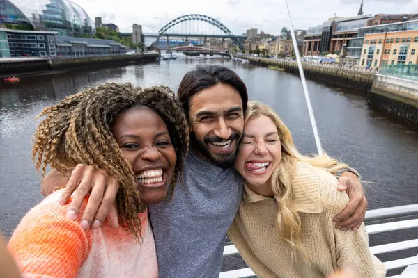 İngiltere, Newcastle 'da bir köprüden geçerken şehir merkezinde duran üç orta yaşlı arkadaşın selfie çekimi. Günlük giysiler giyiyorlar ve yan yana duruyorlar, gülümsüyorlar ve aptal bir grup selfie 'si çekerken kameraya bakıyorlar..