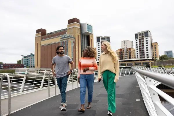 Üç yetişkin arkadaşın Newcastle, İngiltere 'de bir köprüden geçerken bir şehir caddesinde yürürken ön görüntüsü. Günlük giysiler giyiyorlar ve yan yana yürüyorlar, gülümsüyorlar ve sohbet ediyorlar..