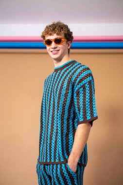 Kameraya gülümseyen, elleri cebinde güneş gözlüğü takan genç bir adamın dörtte üçlük bir stüdyo görüntüsü. Kahverengi kıvırcık saçları var ve mavi ve kahverengi çizgili örgülü bir elbise giyiyor..