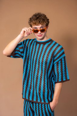 Üç çeyrek boyunda bir stüdyo çekiminde yetişkin bir erkek kameraya tarafsız bir ifadeyle bakıyor. Güneş gözlüğü takıyor, mavi ve kahverengi çizgili örgü örüyor. Bir eli güneş gözlüğünü tutarken cebinde. 
