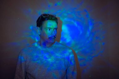 Yaratıcı bir stüdyo resmi. Bıyıklı, genç, yetişkin bir erkek. Tarafsız bir ifade takınıyor. Neon renkli ışıklar onun üzerinde parıldıyor..