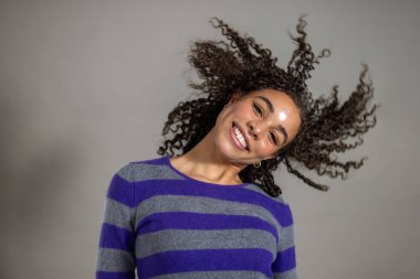 Kıvırcık saçlı, gülümseyen ve yüzünü kameraya doğru bükülmüş bir şekilde çeviren genç bir kadının stüdyo fotoğrafı. Çizgili bir kazak giyiyor, gri bir sırt çantasına karşı duruyor..