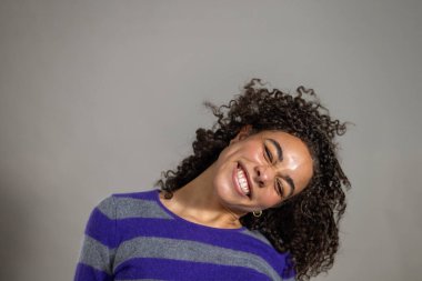 Kıvırcık saçlı, gülümseyen ve yüzünü kameraya doğru bükülmüş bir şekilde çeviren genç bir kadının stüdyo fotoğrafı. Çizgili bir kazak giyiyor, gri bir sırt çantasına karşı duruyor..