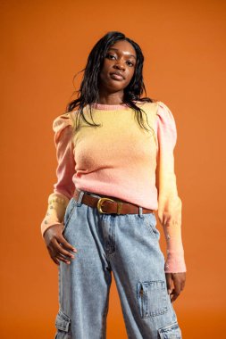 Kameraya ciddi bir ifadeyle bakan genç bir kadının stüdyo resminin dörtte üçü. Günlük kıyafetler giyiyor, turuncu bir stüdyonun arka planında duruyor..