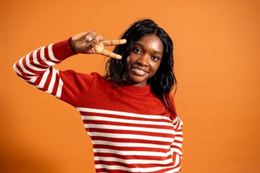 Kameraya gülümseyen, tek eliyle barış işareti yapan genç yetişkin bir kadının belden yukarı stüdyo resmi. Çizgili kazak giyiyor, turuncu bir stüdyonun arka planında duruyor..