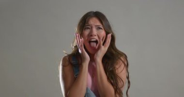 Genç yetişkin bir kadının kameraya iki eliyle korkunç bir yüz ifadesi gösteren video klibi. Uzun esmer saçları var ve altında soluk pembe bir atlet olan kot tulumlar giyiyor..