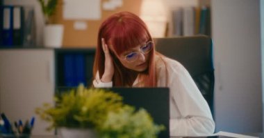 Ofiste dizüstü bilgisayar kullanırken buruşan kâğıttan endişelenen kızıl saçlı genç iş kadını..