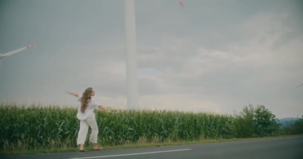 在风电场附近的路上散步的无忧无虑的年轻女子慢镜头 — 图库视频影像