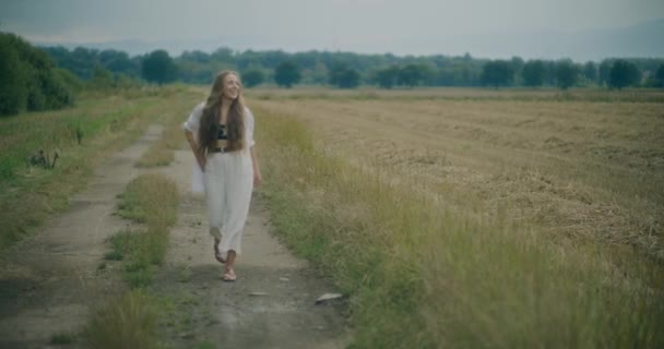 一个微笑的年轻女子 双手插在口袋里 在田野的小径上与天空相对 慢镜头 — 图库视频影像