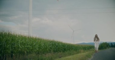Yavaş çekim, elleri ceplerinde genç bir kadın rüzgar çiftliği yakınlarında gökyüzüne karşı yürüyor.