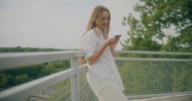 Yavaş çekim, gülümseyen kadının gökyüzüne yaslanmış korkuluklara yaslanırken telefonla mesajlaşması.