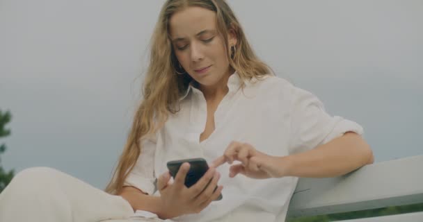 Sarışın Kadının Gökyüzüne Karşı Bankta Otururken Akıllı Telefonu Kaydırdığı Yavaş — Stok video
