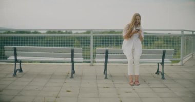 Sarışın kadının cep telefonu kullanırken, korkulukların yanındaki bankta gökyüzüne yaslanırken yavaş çekim.