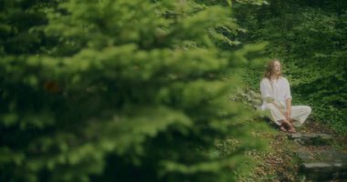 Genç bir kadının yeşil ormandaki bitkilerin yanında otururken taze hava soluduğu yavaş çekim.