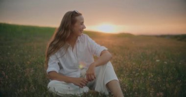 Gün batımında çiçeklerin arasında gökyüzüne karşı otururken genç ve mutlu bir kadının yavaş çekimleri.